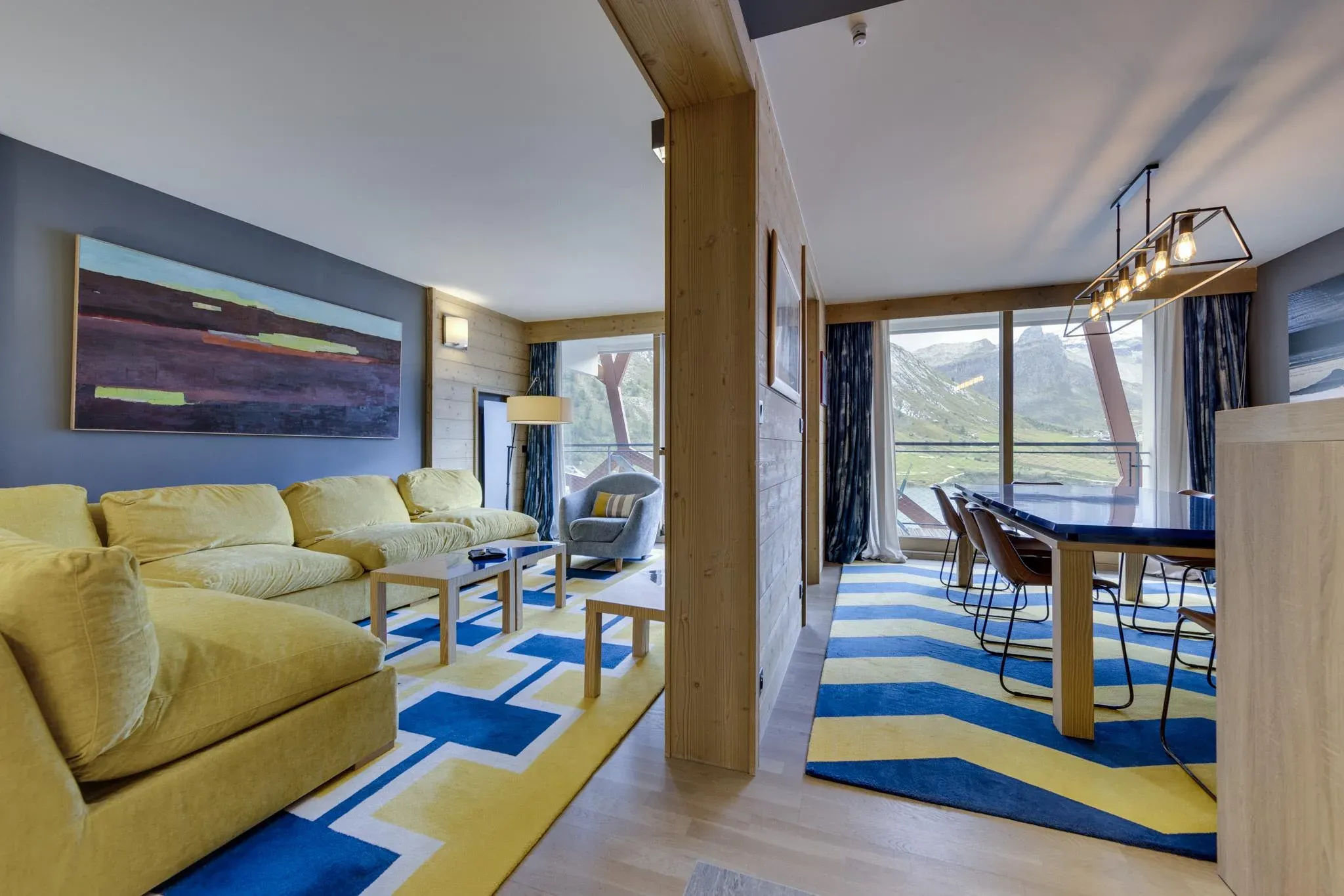 Open-plan lounge with yellow sofa, coffee tables and dining table beside mountain-view windows